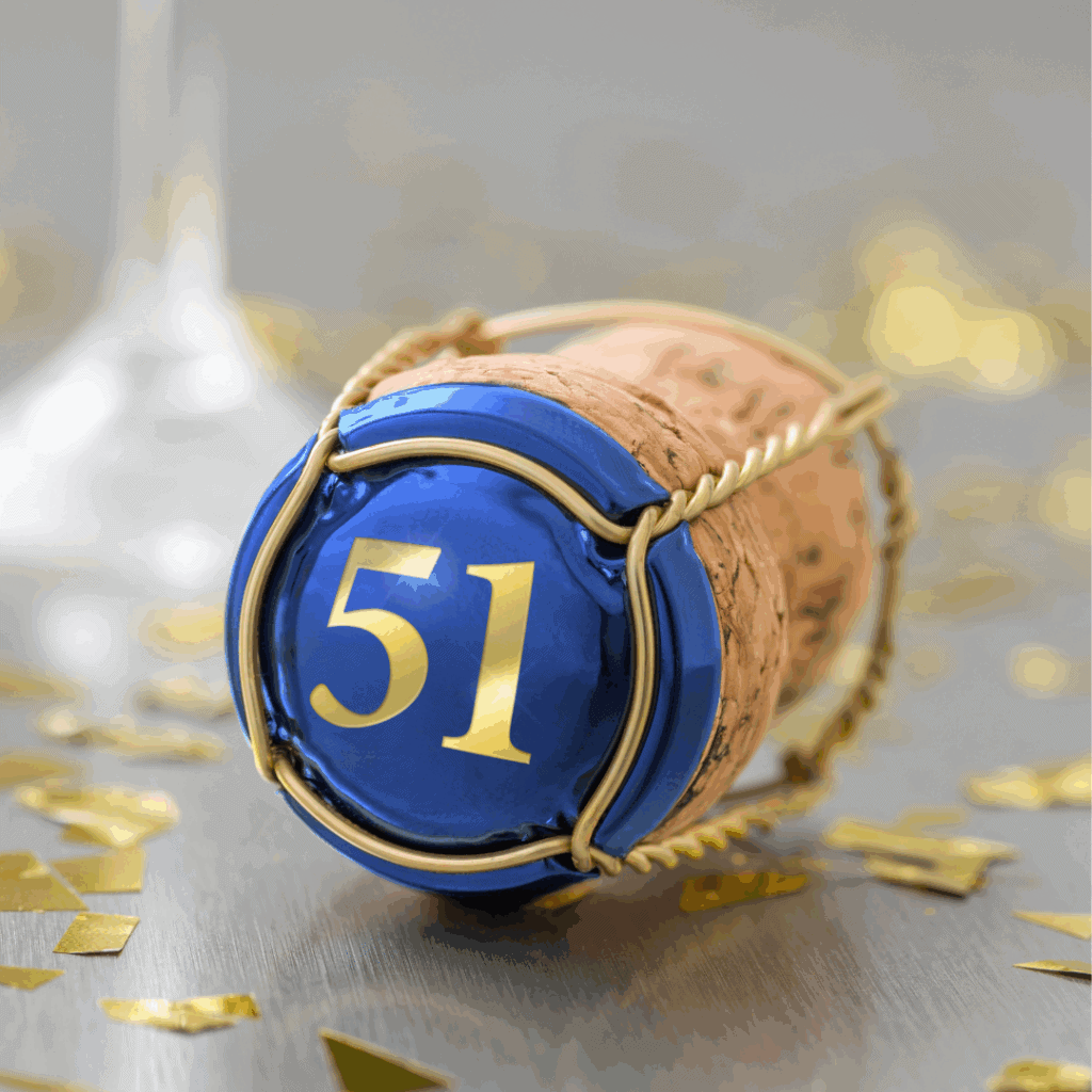 A close-up of a champagne cork with a metallic blue cap labeled “51,” surrounded by gold confetti—celebrating a milestone with intention and reflection.