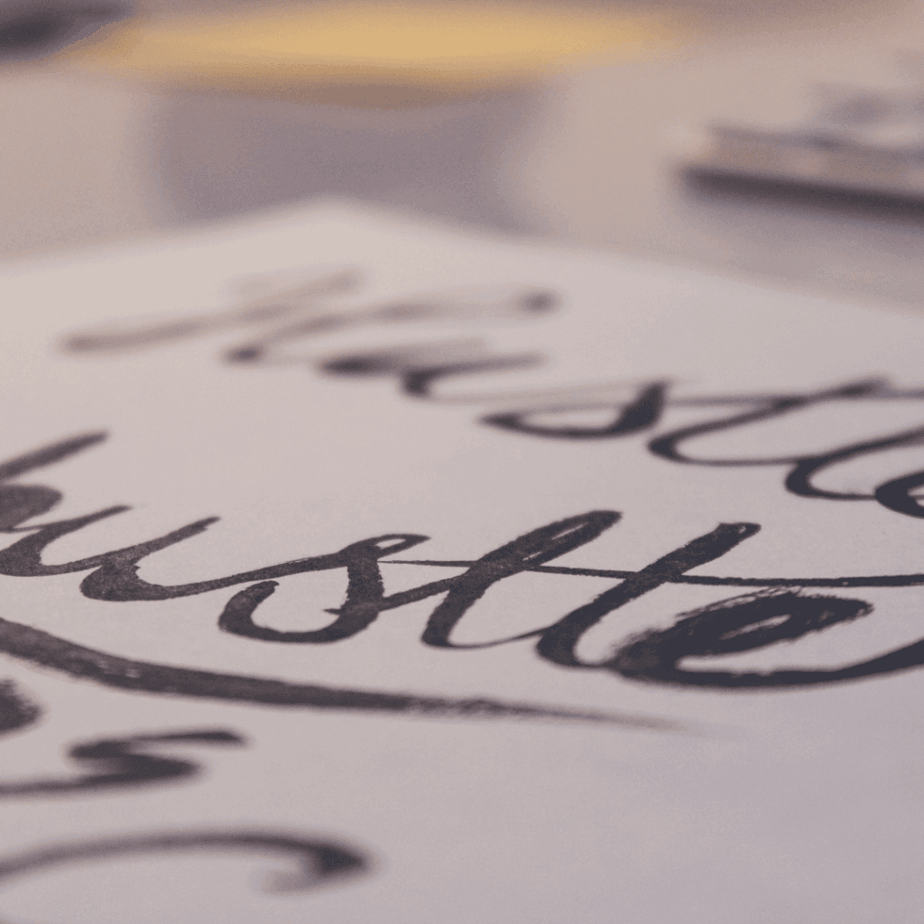 Close-up of the word “hustle” written in bold, artistic script on white paper, with a blurred background suggesting motion and focus.