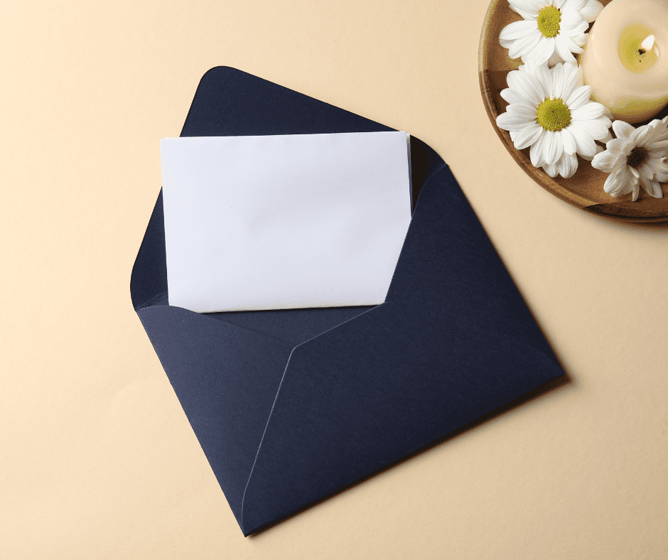 A handwritten letter partially tucked into a dark envelope, resting on a soft surface beside a lit candle and white flowers.