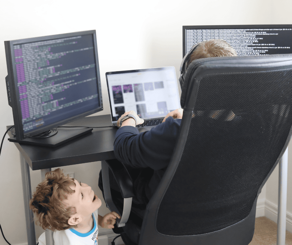 Work-from-home dad at a desk while his young child looks up from underneath, illustrating the overlap of remote work and family life.