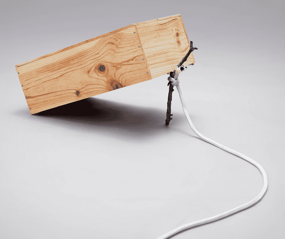A wooden box propped up by a stick with a string attached, staged as a primitive trap — a visual metaphor for the deceptive appeal of "trying," which appears to be action but collapses the moment it's tested.
