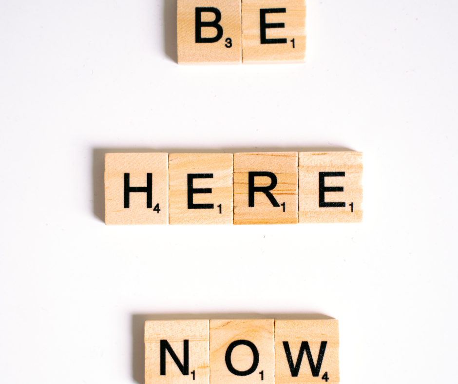 The phrase BE HERE NOW spelled out in wooden blocks and spaced separately on a white background, emphasizing the HERE as the place between BE and NOW. All 3 are are locations more than anything else.