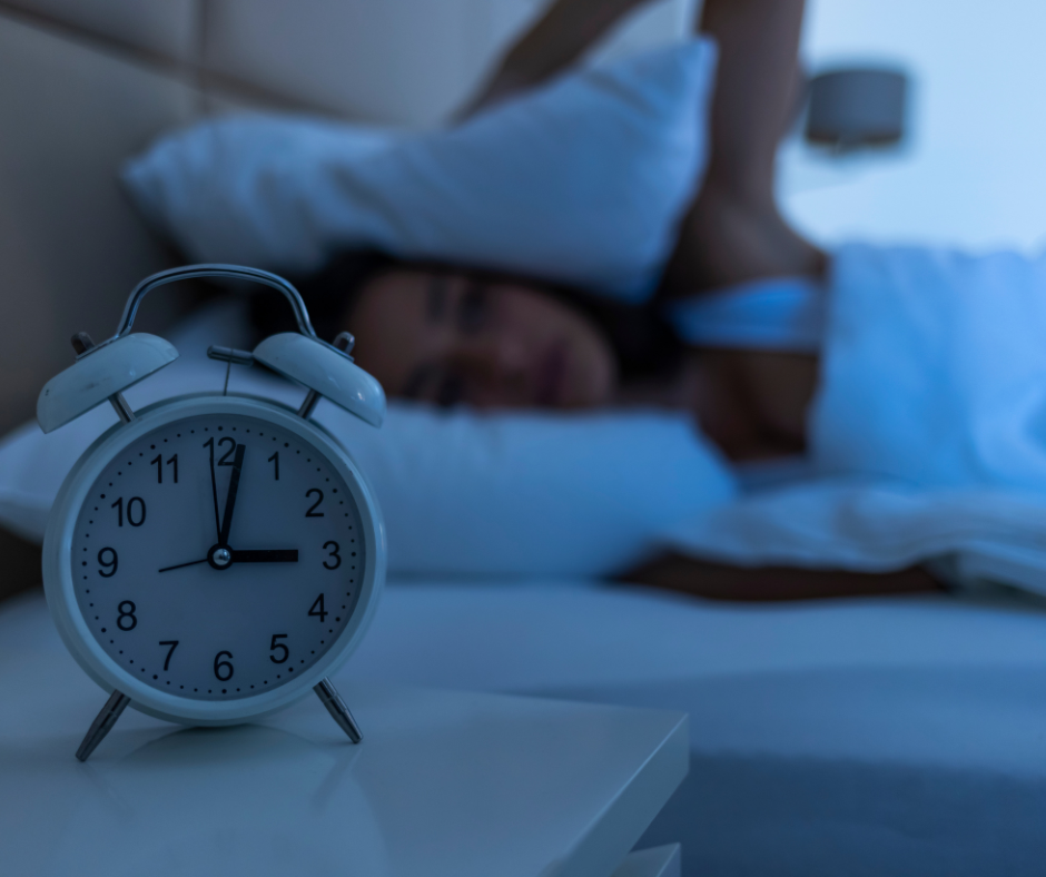 A white twin-bell alarm clock reads about 3:00 on a nightstand, sharply in focus, while a person lies awake in the blue-lit background, covering their head with a pillow — the frustration of trying too hard to sleep.