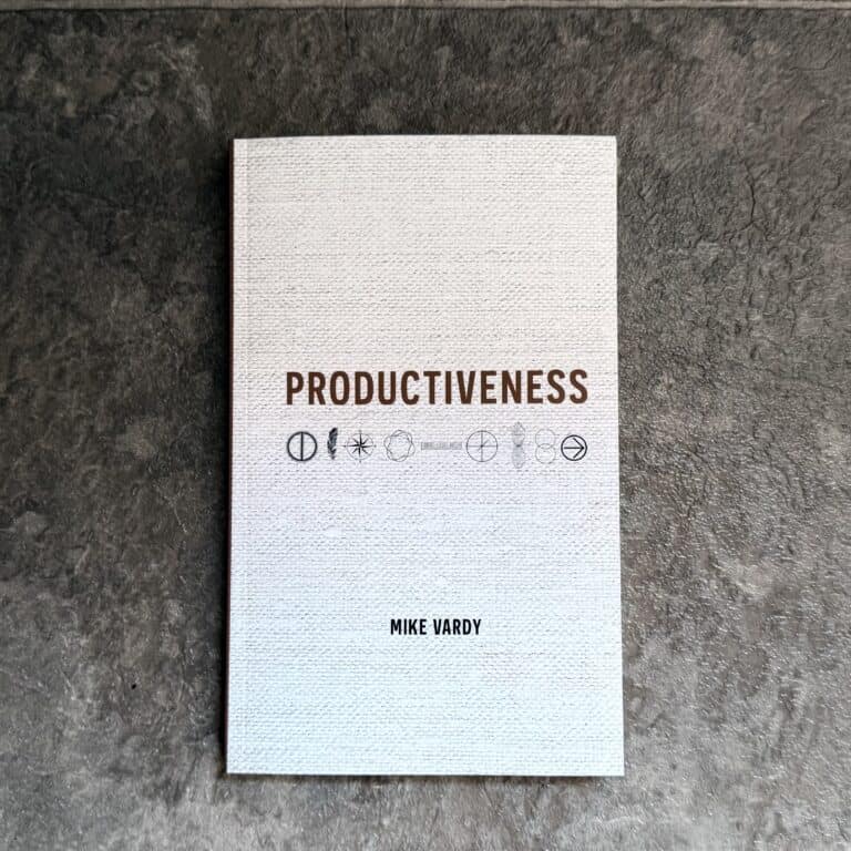 Paperback copy of Productiveness by Mike Vardy on a dark concrete surface — the title in brown above a row of nine hand-drawn principle symbols on a linen-textured white cover